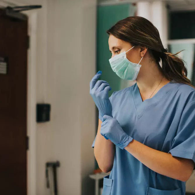 Nurse in blue scrubs and mask putting on gloves in a hospital room.